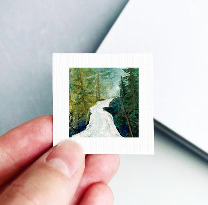 A hand is holding a small square artwork depicting a winding river surrounded by lush green trees.