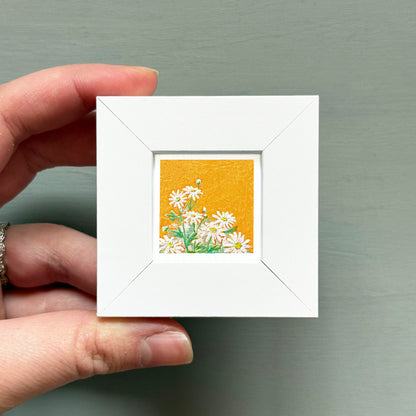 A hand is holding a small white frame containing a painting of white flowers against an orange background.