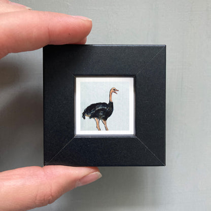 A person's hand is holding a small black square frame containing a picture of an ostrich.