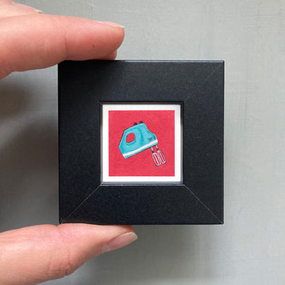 A hand is holding a small black square frame containing a vibrant red square with a blue and white illustration of a hand mixer.