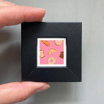 A person's hand holding a small square frame containing an illustration of various pastries, including a croissant, a donut, and a muffin.