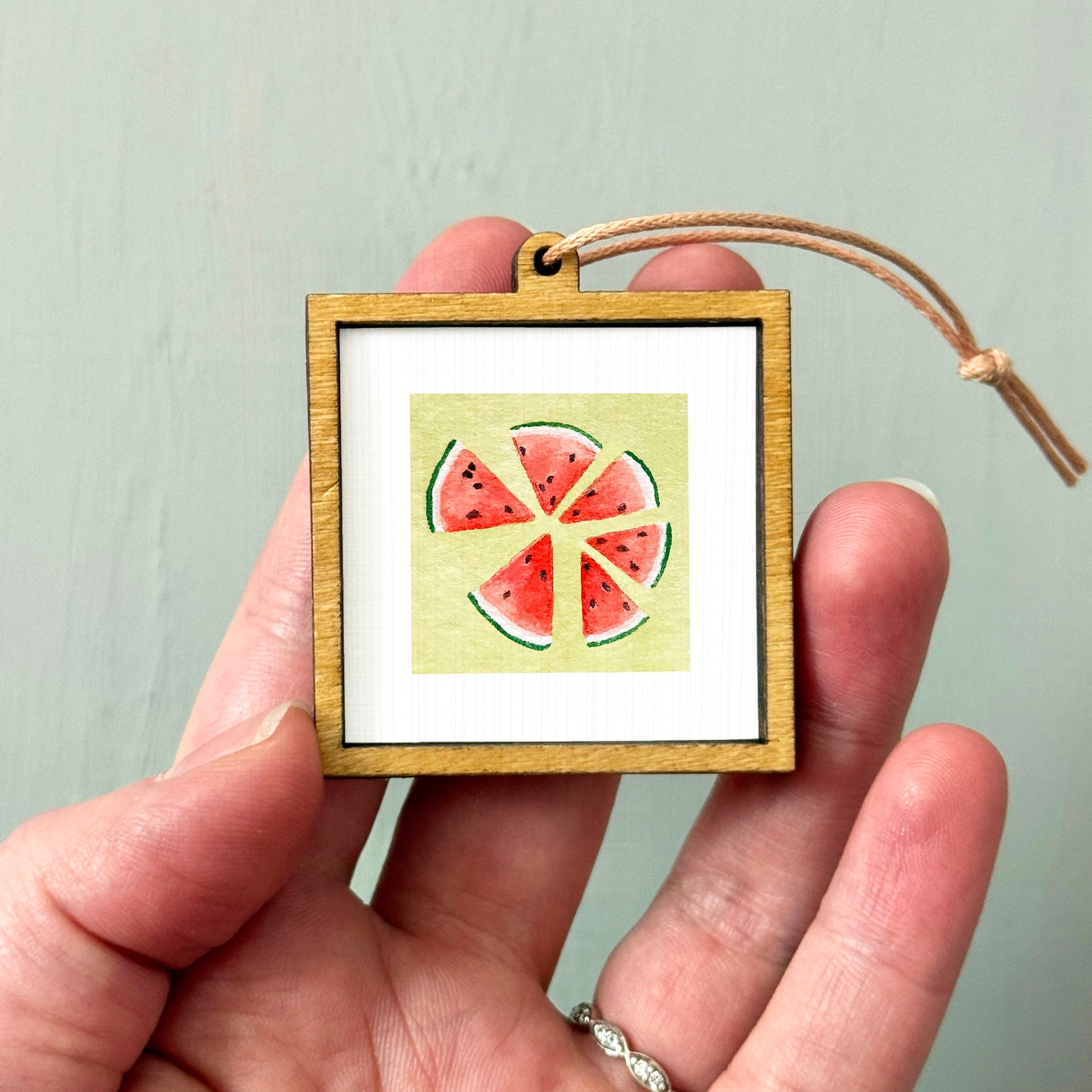 A hand holding a small wooden frame containing a watermelon slice illustration.