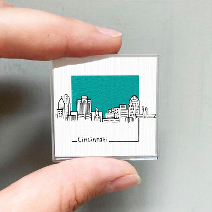 A hand holding a small square magnet with a cityscape illustration on it, featuring a teal background and the word "CINCINNATI" written below.