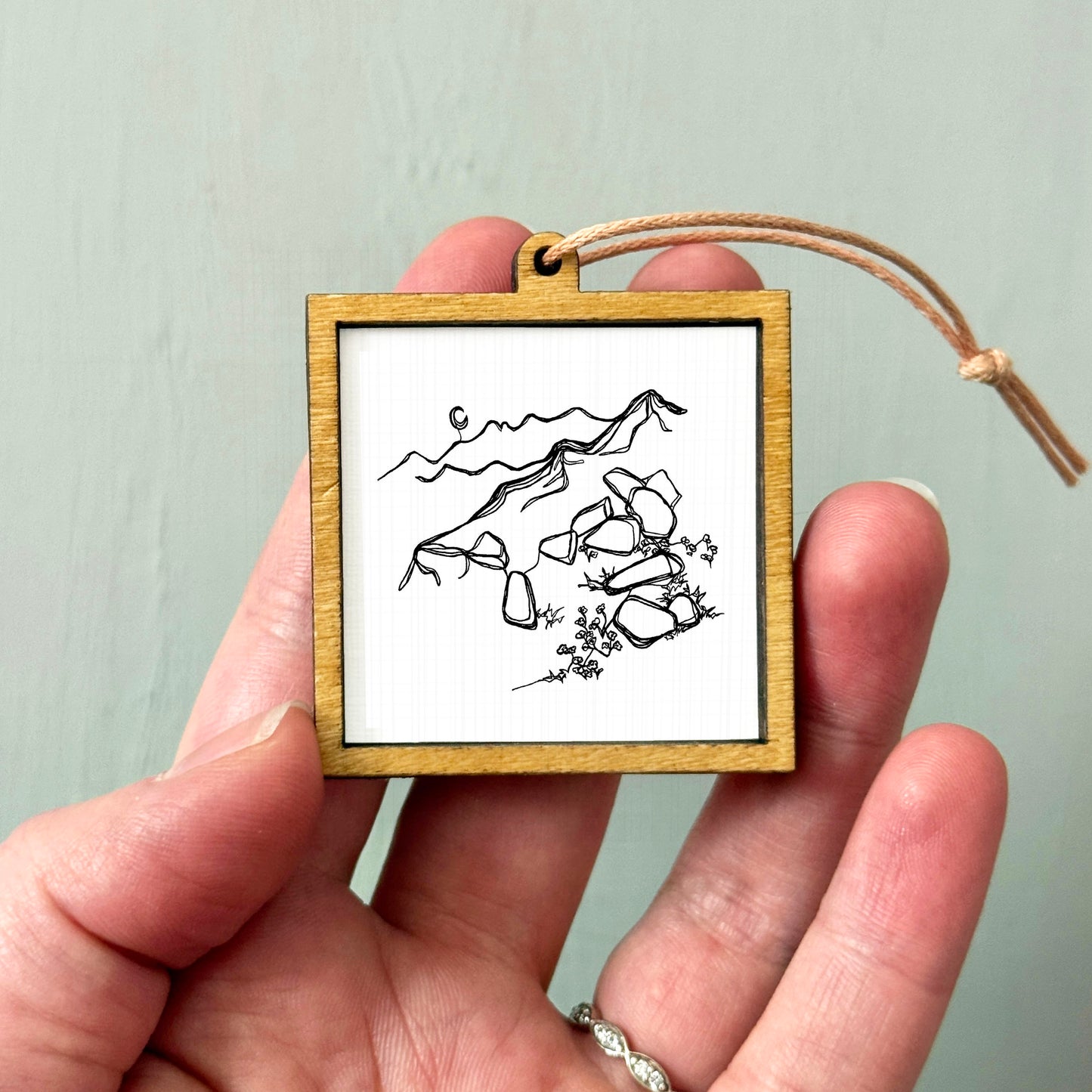A hand holding a small wooden frame containing a black and white line drawing of mountains and rocks.