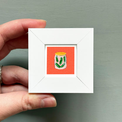 A hand is holding a small white square frame containing an illustration of a jar with green pickles on an orange background.