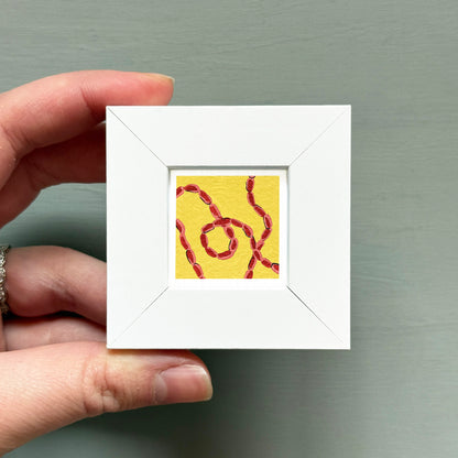 A hand is holding a small white square frame containing a vibrant yellow and red abstract painting.