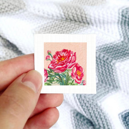 A hand is holding a small square card with a painting of pink flowers on it, against a background of a white and gray striped blanket.