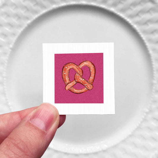 A hand is holding a small square card with an illustration of a pretzel on it.