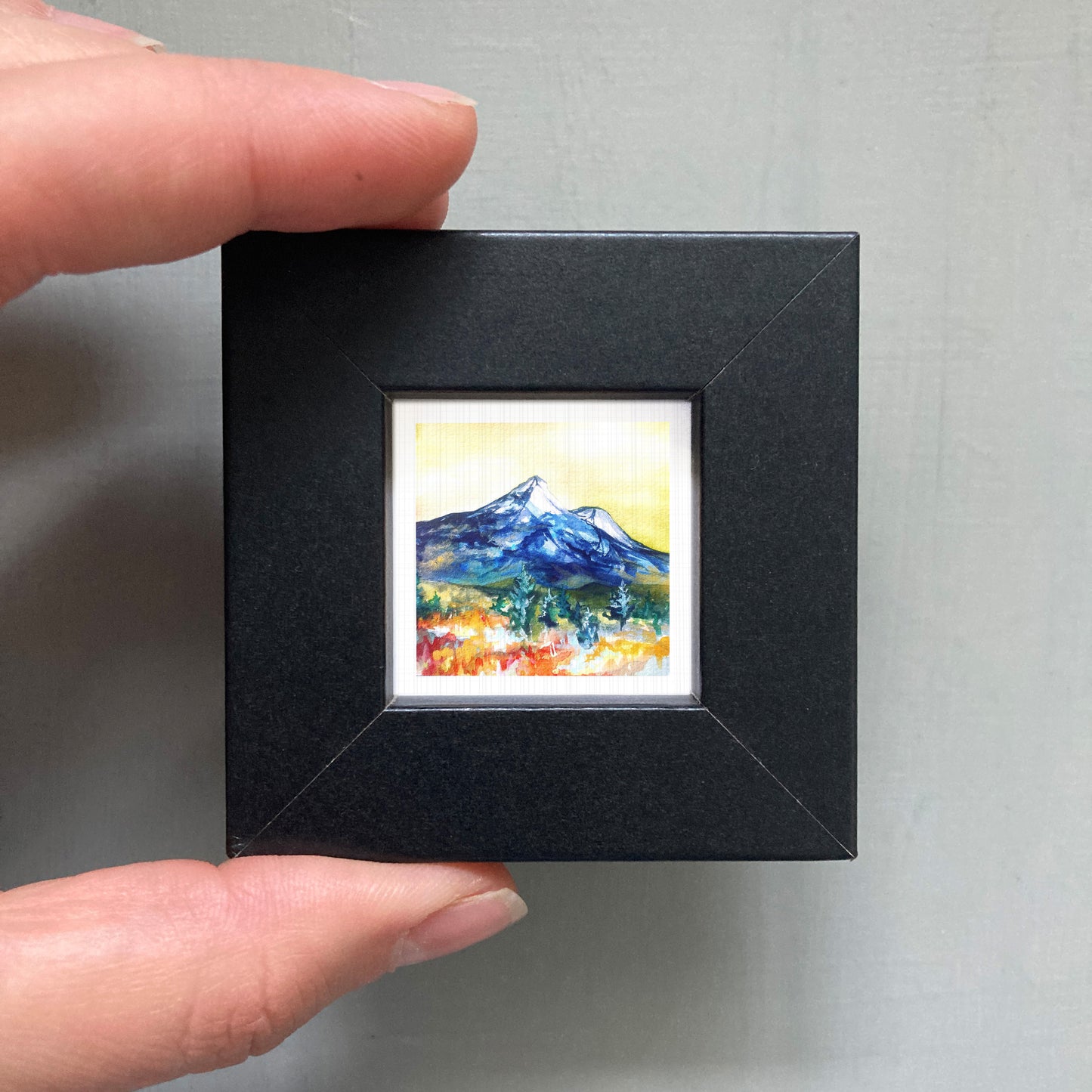 A person's hand is holding a small, square-shaped black frame containing a vibrant, colorful painting of a mountain landscape.