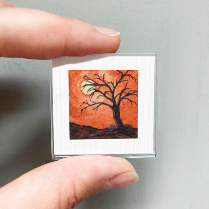 A person's hand is holding a small square picture depicting a tree with a full moon in the background.