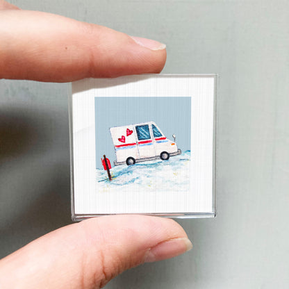 A person's hand is holding a small square picture depicting a white ambulance with red hearts on its side, standing on a snowy surface.