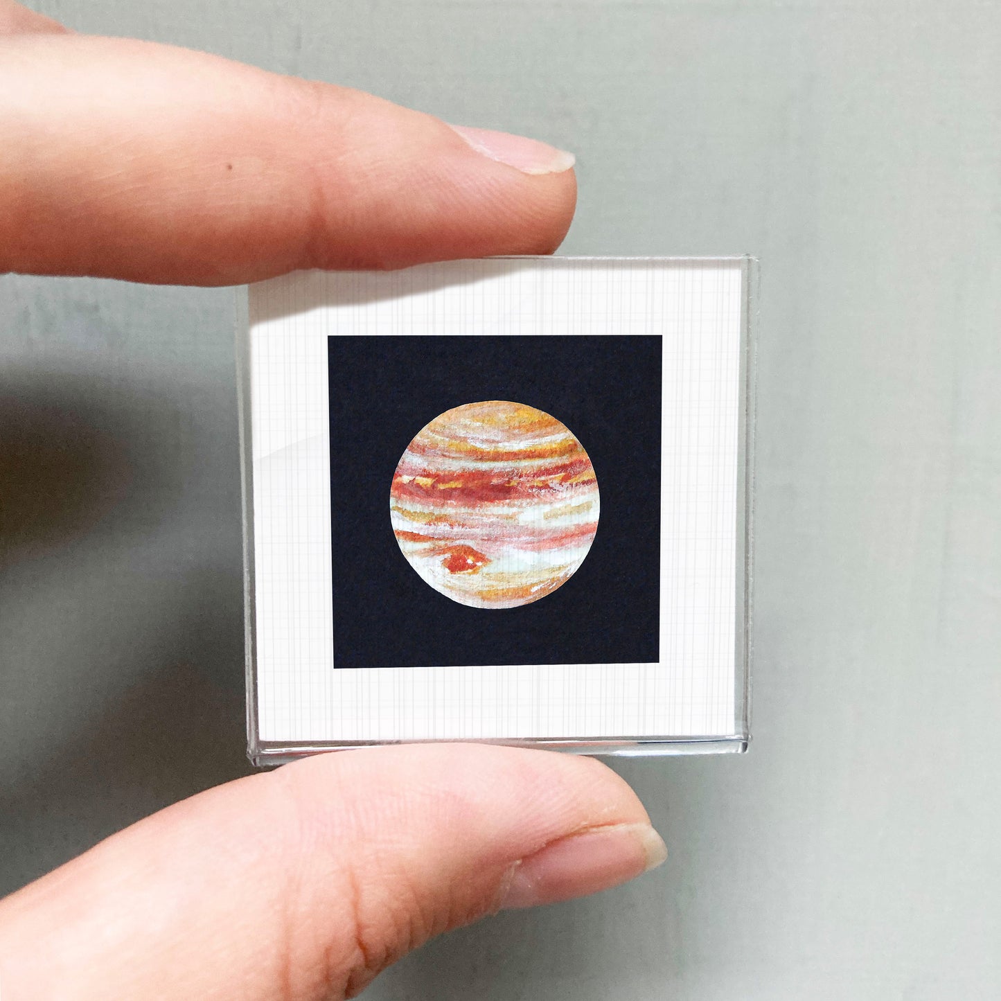 A person's hand is holding a small square frame containing a detailed image of the planet Jupiter.