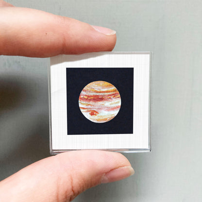 A person's hand is holding a small square frame containing a detailed image of the planet Jupiter.