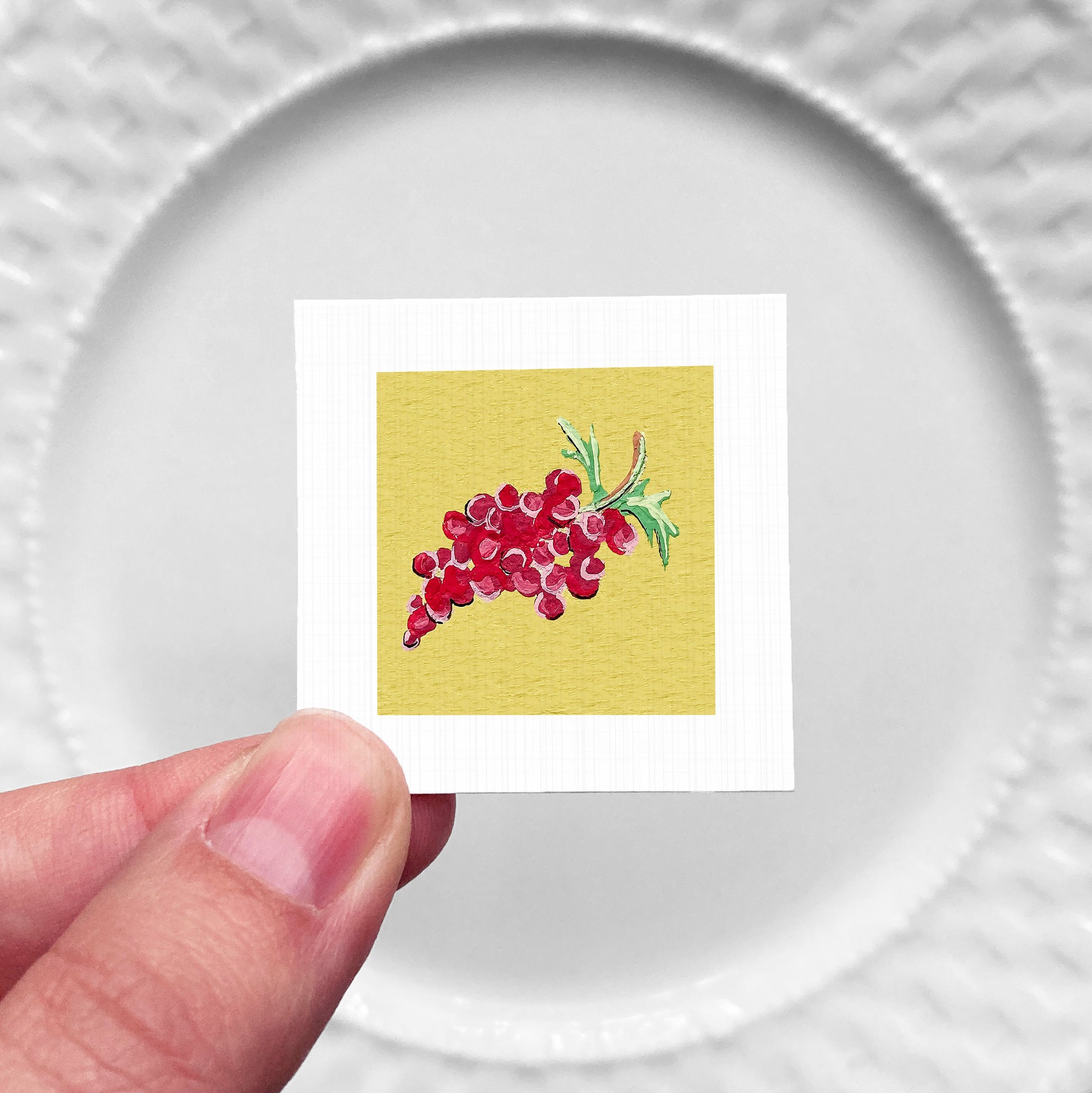 A hand is holding a small square card with a vibrant illustration of red grapes on a yellow background.