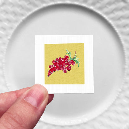 A hand is holding a small square card with a vibrant illustration of red grapes on a yellow background.
