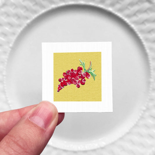 A hand is holding a small square card with a vibrant illustration of red grapes on a yellow background.