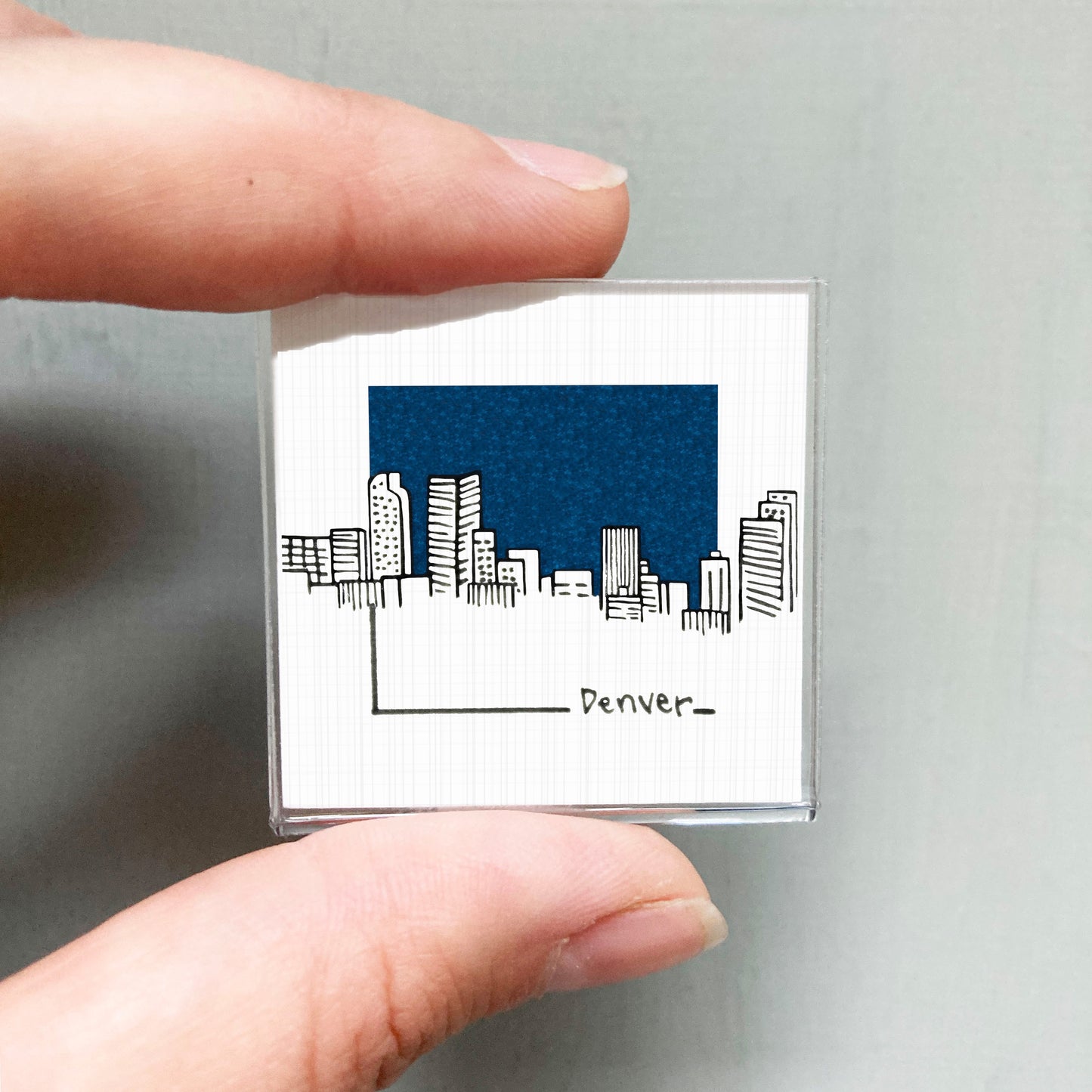 A hand holding a small square magnet with a drawing of a city skyline and the word "Denver" written below it.