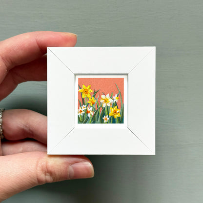 A hand is holding a small white square frame containing a painting of yellow and white flowers.