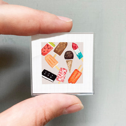 A person's hand holding a small square with various colorful ice cream cones and popsicles depicted on it.