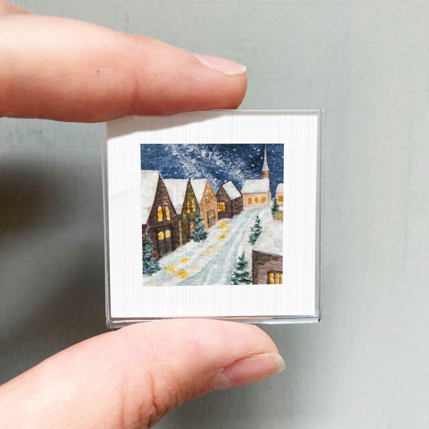 A hand holding a small square picture depicting a snowy village scene with houses and trees.