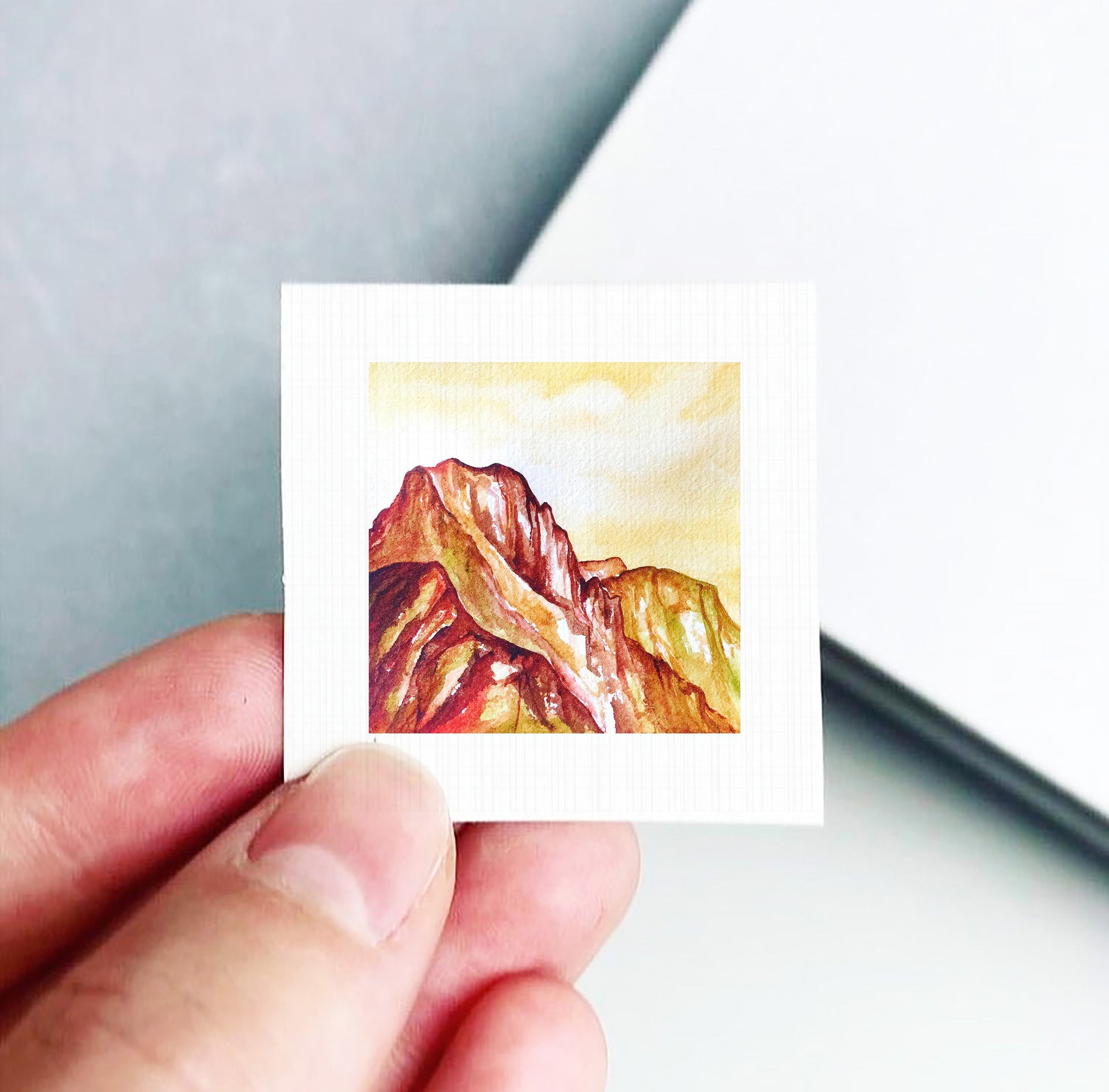 A hand is holding a small square card with a painting of a mountain landscape on it.