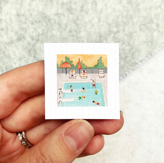 A hand holding a small square picture depicting a swimming pool with people enjoying the water and lounging on chairs.
