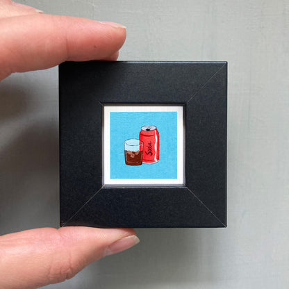 A person's hand holding a small black square frame containing an illustration of a red can of soda and a glass of soda.