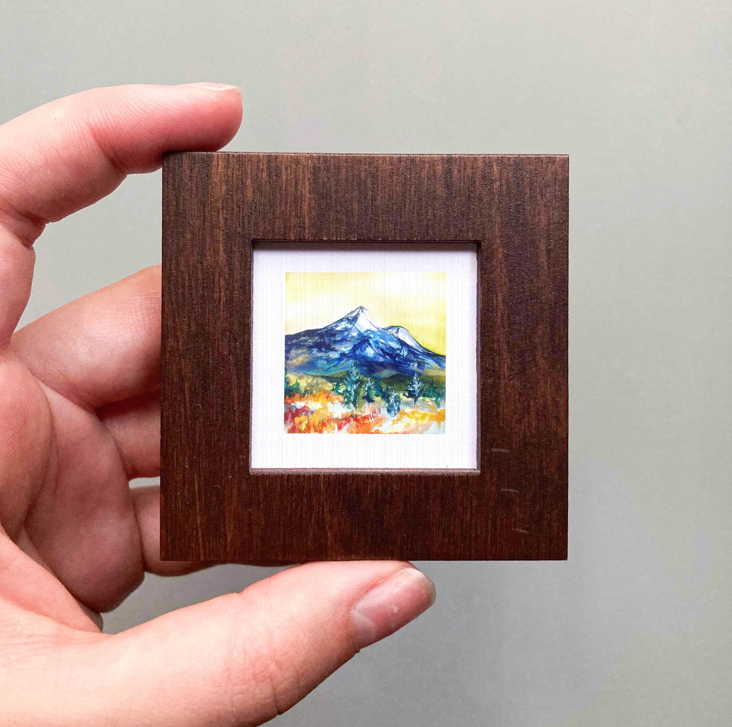 A hand is holding a small wooden frame containing a watercolor painting of a mountain landscape.
