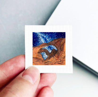 A hand is holding a small square-shaped piece of paper with a painting of a desert landscape with a starry sky on it.