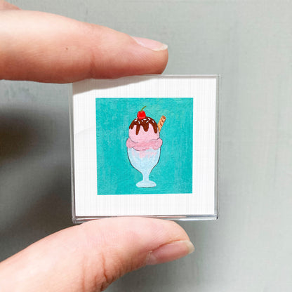 A person's hand holding a small square picture of a pink ice cream cone with a cherry on top.