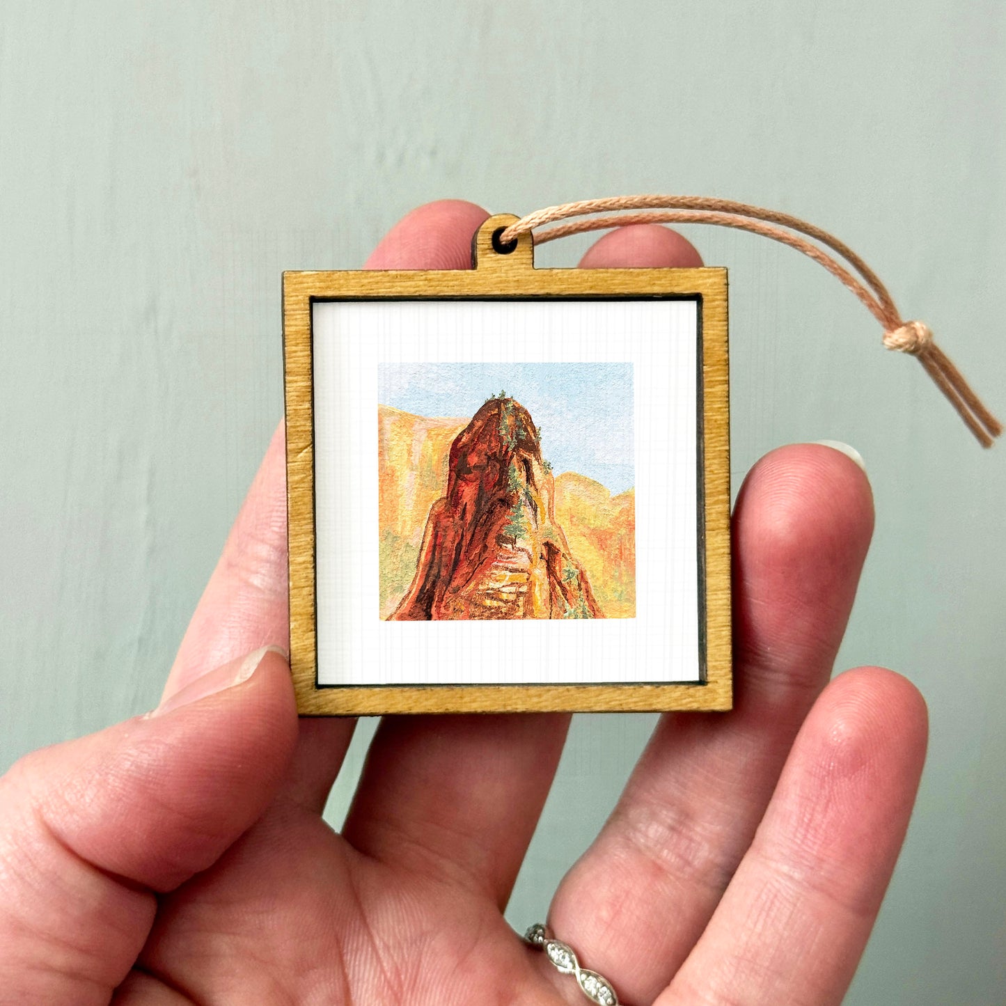 A person's hand holding a small wooden frame containing a framed painting of a mountain landscape.